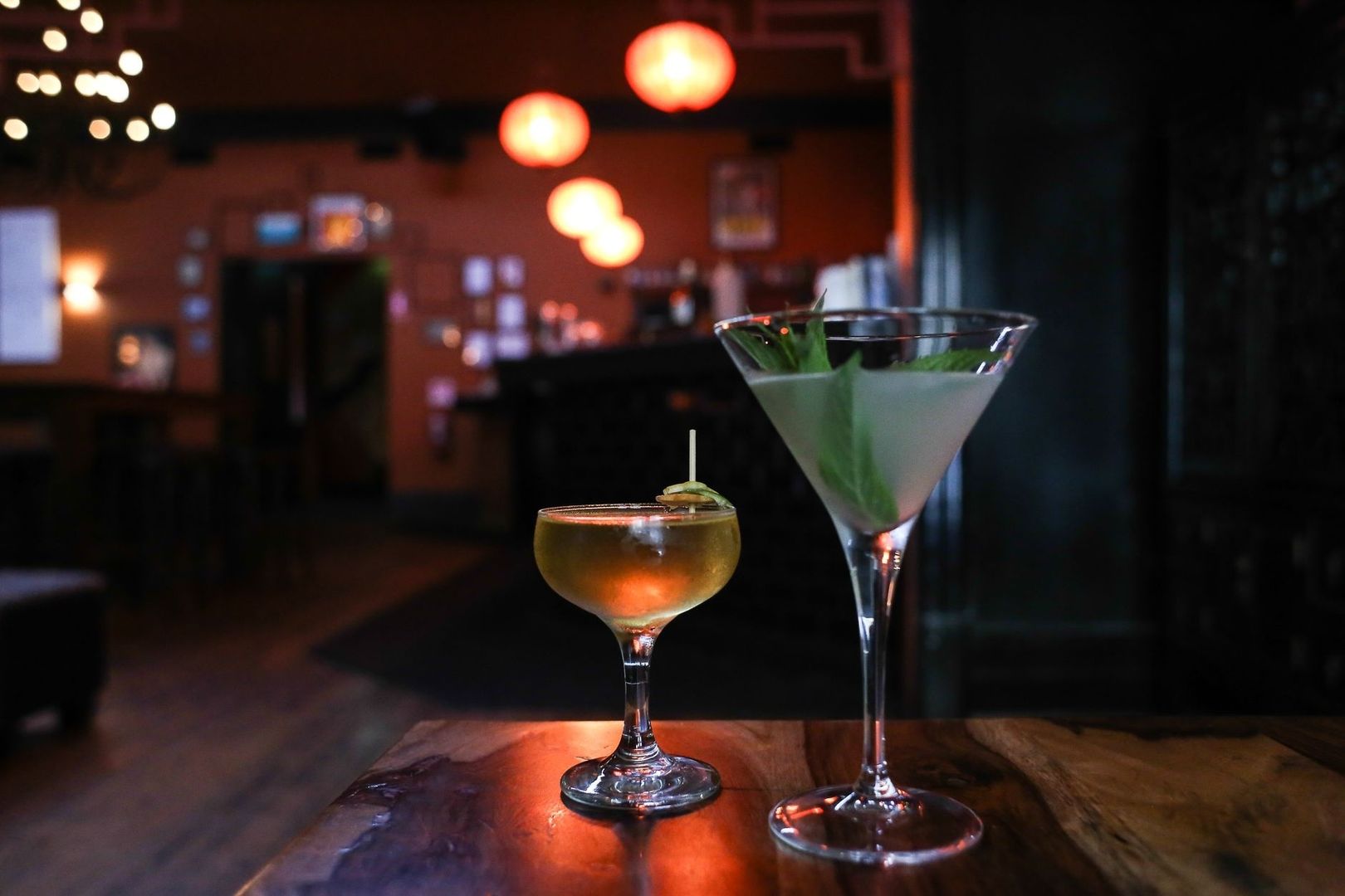 Two cocktails on a wooden table in a warmly lit, cozy bar
