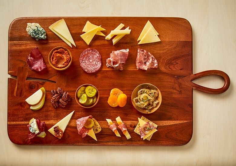 Wooden board arranged with Swiss cheeses, cured meats, fruit, and pickles